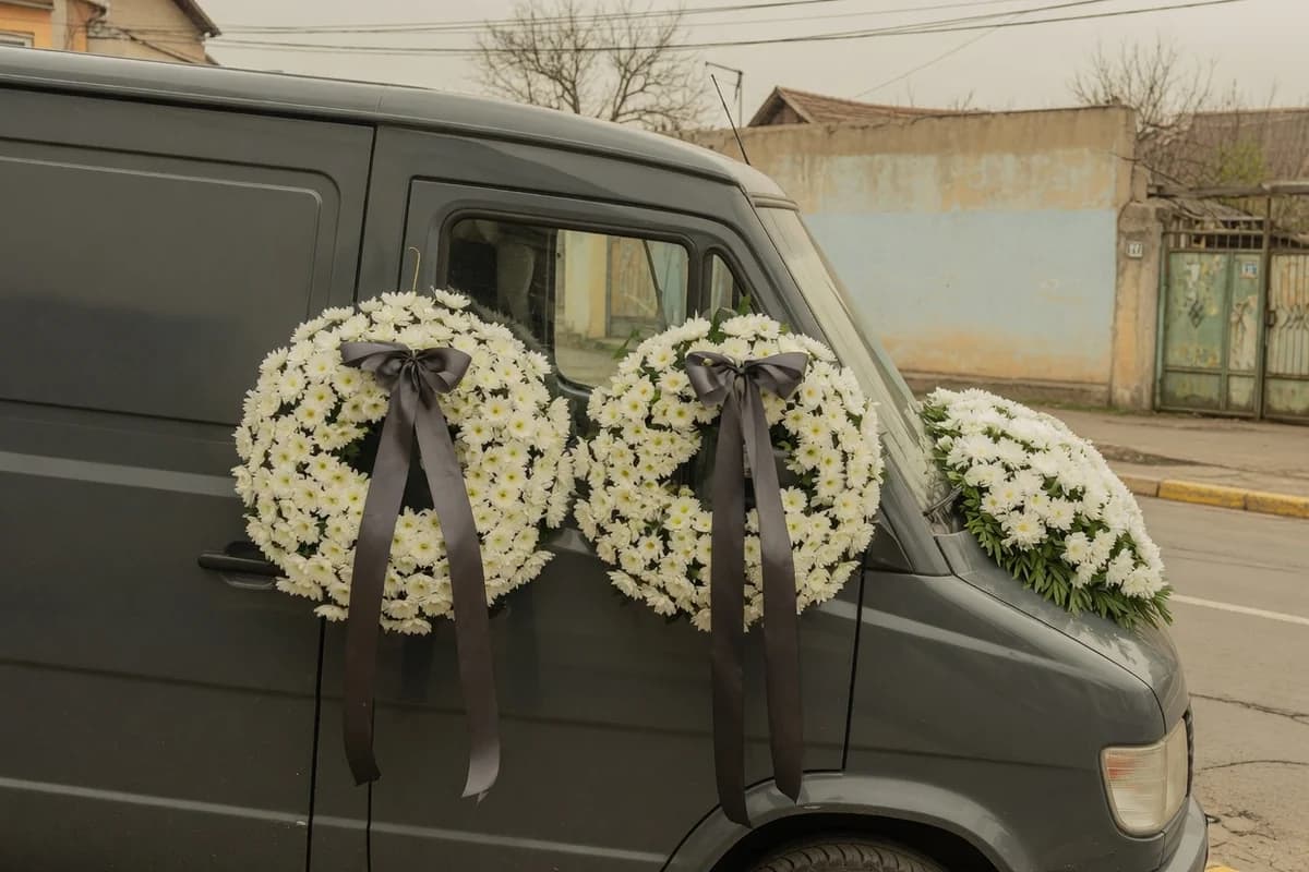 Aranjamente florale funerare fixate pe capota și laterala unui vehicul funerar, pregătite de cortegiu